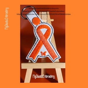 Pode incluir: Fita laranja com uma borboleta, bordada em um fundo branco. A fita está presa a um chaveiro. A fita é um símbolo de conscientização para múltiplas causas, incluindo leucemia, esclerose múltipla e câncer de rim.