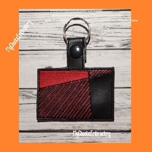 May include: A black and red plaid key chain with a black leather strap and a silver key ring. The key chain is embroidered with a plaid pattern and has a black leather backing. The text "MyChaoticEmbroidery" is printed on the key chain.