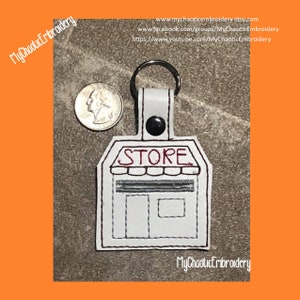 Op de afbeelding: Witte leren sleutelhanger met een rood geborduurd winkelbord met de tekst "STORE" en een zwarte knop. De sleutelhanger is ongeveer zo groot als een kwartje.