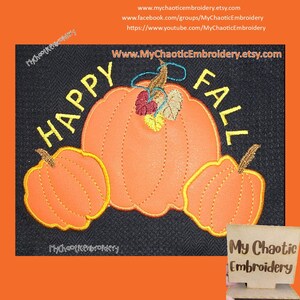 Op de afbeelding: Borduurwerk met de tekst "Happy Fall" met drie oranje pompoenen en herfstbladeren. Het ontwerp is geborduurd op een zwarte stoffen achtergrond.
