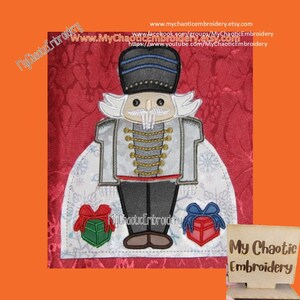 May include: Embroidered design of a Nutcracker holding a gift. The Nutcracker is wearing a gray coat with gold buttons, a black hat, and a white beard. The design is on a red background with snowflakes.