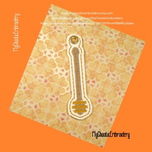 May include: An embroidered honey dipper with gold glitter detailing and a white border. The dipper is set against a patterned background. The text "MyChaoticEmbroidery" and website details are visible.