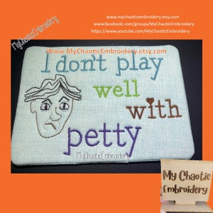 May include: A light blue embroidered fabric coaster with the text "I don't play well with petty" and a cartoon face with a frown. The text is in brown, green, and purple thread. The coaster is on a bright orange surface.
