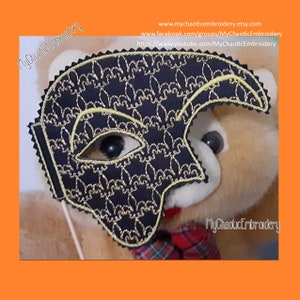May include: A black and gold masquerade mask with a fleur-de-lis pattern. The mask is held up by a stick and is being worn by a teddy bear.