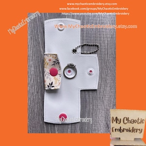 May include: White leather ring holder with a floral design and a pink button. The holder has a small metal ring attached to it. The text "My Chaotic Embroidery" is printed on the holder.