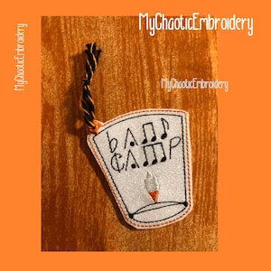 May include: White felt ornament shaped like a drum with the text "Band Camp" and musical notes embroidered in black thread. The ornament has a black and orange string attached to the top.