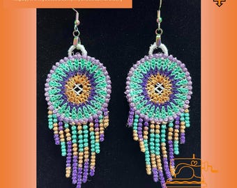 Peut inclure: Une paire de boucles d'oreilles faites à la main avec un motif rond brodé multicolore et une frange de perles. Les boucles d'oreilles sont vertes, violettes, dorées et blanches.