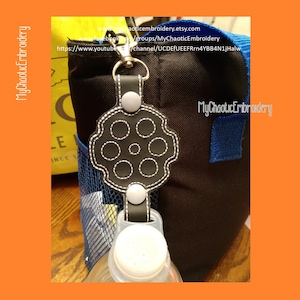 以下が含まれることがあります： 白いステッチで銃身のデザインが施された黒い革製のボトルホルダー。ホルダーの両側には白いボタン、上部には金属製のリングが付いています。