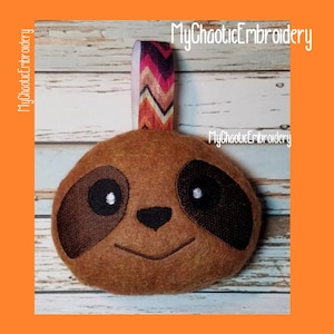 May include: A brown felt sloth ornament with embroidered eyes, nose, and mouth. The ornament has a brown and orange striped ribbon attached to the top.