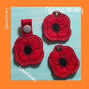 May include: Three red felt poppy flower keychains with black centers. The keychains have white stitching around the edges. One keychain has a silver button on the top. The keychains are on a light blue background. MyChaoticEmbroidery is written on the image.