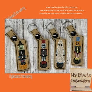 May include: Four felt Halloween keychains with different characters: a witch, a skeleton, a ghost, and a pumpkin. Each keychain is attached to a brown felt rectangle with a black button and a silver key ring. The keychains are on a wooden surface. The text "My Chaotic Embroidery" is visible in the bottom right corner of the image.
