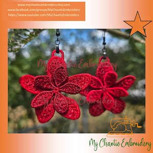 May include: A pair of red flower earrings. Each earring features a detailed, embroidered flower design with five petals. The earrings hang from black hooks. The background is blurred, with green foliage visible.