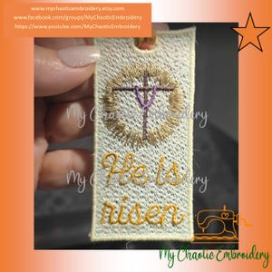 May include: Embroidered rectangular tag with the text "He is risen" in gold thread. The tag features a gold wreath with a purple cross at the center. The background is a light cream color. The tag is held by a person.