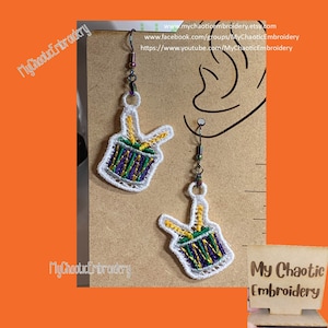 May include: A pair of white embroidered earrings with a purple, green, and gold drum design. The earrings are hanging from silver hooks. The text "My Chaotic Embroidery" is visible in the image.