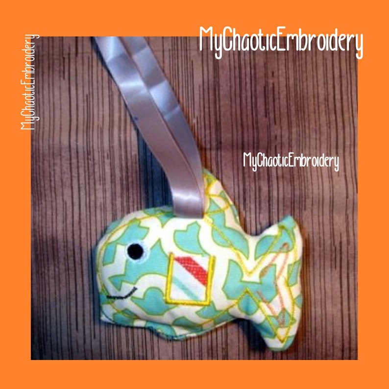 May include: A teal and yellow patterned fish-shaped stuffed toy with a black eye and a red and white patterned square on its side. The toy is hanging from a grey ribbon and the text "MyChaoticEmbroidery" is visible in the background.
