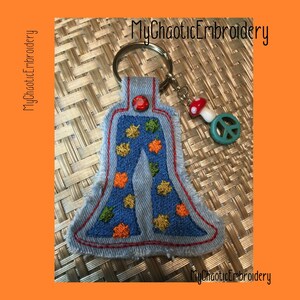 以下が含まれることがあります： 青いデニムのキーホルダーに、ベルボトムのズボンが刺繍されています。ズボンには、オレンジ、イエロー、グリーンの葉が刺繍されています。キーホルダーには、赤と白のキノコのチャームと、緑の平和のサインのチャームも付いています。
