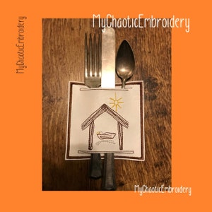 May include: A white fabric napkin with an embroidered image of a manger scene. The napkin is holding a fork, knife, and spoon. The text "MyChaoticEmbroidery" is visible in the top right corner and bottom right corner of the image.