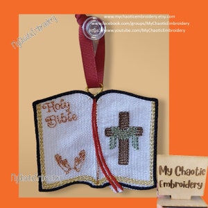 May include: A white embroidered ornament shaped like an open book with the words "Holy Bible" on the left page and a brown cross with green leaves on the right page. The ornament has a red ribbon attached to the top.