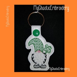 May include: White leather keychain with a green shamrock gnome design. The gnome is wearing a green hat and holding a shamrock. The keychain has a metal ring for attaching keys.