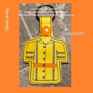 Könnte beinhalten: Gelber bestickter Schlüsselanhänger in Form eines Hemdes mit einem schwarzen Knopf und einem orangefarbenen Gürtel. Der Schlüsselanhänger hat einen silbernen Ring und einen kleinen orangefarbenen Knopf.