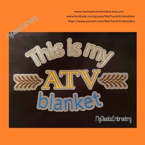 May include: Black embroidered design on a dark fabric with the text "This is my ATV blanket" and a wheat stalk design. The text is in white, yellow, and blue. The wheat stalk is in brown. The background is orange.