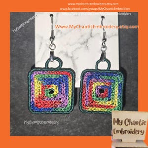 May include: A pair of square, handmade earrings with a colorful, concentric design. The earrings feature a rainbow of colors, including red, yellow, green, blue, and purple, and are outlined in dark green. The earrings are suspended from silver-colored hooks.