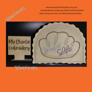 May include: A beige fabric bread of life design with brown embroidery. The design is on a beige fabric with a scalloped edge. The words "Bread of Life" are embroidered in purple and blue thread. The design is on a wooden stand with the words "My Chaotic Embroidery" on it.