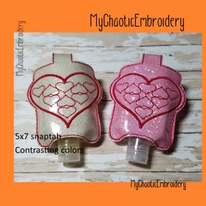 Puede incluir: Dos porta-botellas de desinfectante de manos con un diseño de corazón. Los porta-botellas están hechos de tela blanca y rosa con costuras rojas. El texto "5x7 snaptab" y "Contrasting colors" es visible en la imagen.