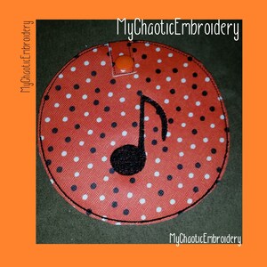 May include: A round, red fabric item with white polka dots and a black musical note design. The item has a black border and an orange snap closure. The text "MyChaoticEmbroidery" is in the corners.