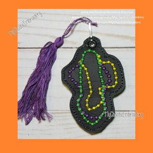 May include: A black leather keychain with a purple tassel. The keychain features a design of Mardi Gras beads in purple, green and gold.