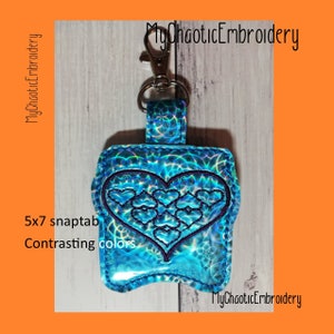 Puede incluir: Un llavero de cierre a presión en forma de corazón azul y morado con acabado holográfico. El llavero mide 5x7 pulgadas y presenta un diseño de múltiples corazones dentro de un corazón más grande. El texto "MyChaoticEmbroidery" es visible en la imagen.