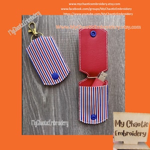 May include: Two red, white, and blue striped fabric keychains with a blue button. One keychain has a gold metal ring and a gold metal clasp. The other keychain has a red fabric pocket with a USB drive inside. The keychain is labeled "My Chaotic Embroidery" on a wooden sign.