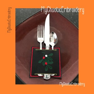 Puede incluir: Un porta cubiertos de tela negra con un diseño de acebo bordado en rojo y verde. El porta cubiertos está sobre un plato marrón y contiene un tenedor, un cuchillo y una cuchara.