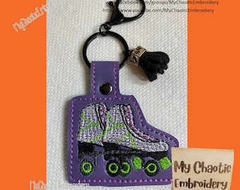 Könnte beinhalten: Lilafarbener Leder-Schlüsselanhänger mit einem grün-weißen Stickmuster von Rollschuhen. Der Schlüsselanhänger hat einen schwarzen Metallring und einen schwarzen Quasten. Der Text "My Chaotic Embroidery" ist auf einem Holzschild im Hintergrund zu sehen.