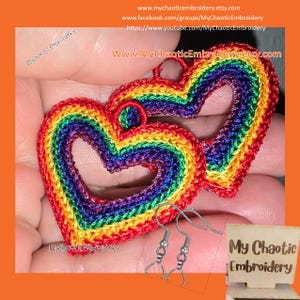 May include: Two rainbow heart-shaped crocheted earrings with silver hooks. The earrings are held in a hand. The image also includes a small wooden sign with the text "My Chaotic Embroidery".