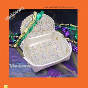 May include: White, scalloped-edge trinket box with gold snap closures and a fleur-de-lis pattern embroidered in gold, green, and purple. The box is open, revealing the interior design. Green, gold, and purple beads are draped around the box.