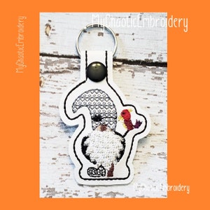May include: White felt gnome keychain with a gray hat and a red and orange turkey. The gnome is holding the turkey in its hand. The keychain has a black button and a silver key ring.