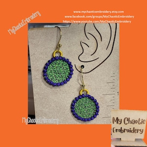 May include: A pair of green and purple embroidered earrings with gold hooks. The earrings are round and have a woven pattern. The earrings are displayed on a brown surface with a ruler.