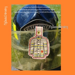 May include: A tan embroidered firefighter jacket keychain with yellow, red, and blue accents. The keychain is hanging from a black firefighter helmet with the word "FIREFIGHTER" on the front. The text "MyChaoticEmbroidery" is visible in the image.