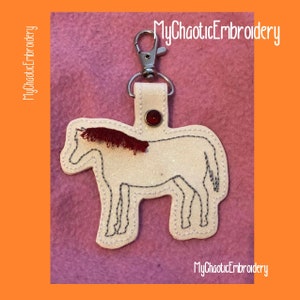 Puede incluir: Llavero de caballo blanco con purpurina, con una crin roja y un botón rojo en la parte superior. El llavero está unido a un anillo de metal plateado.