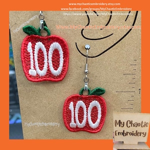 May include: Red apple-shaped earrings embroidered with the number 100 in white. The earrings have green leaf accents and silver-colored hooks. The earrings are displayed on a brown background.