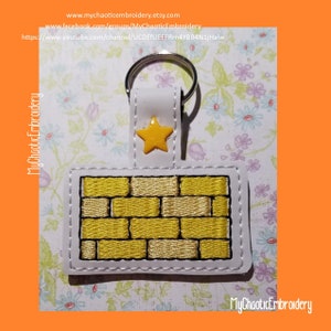 May include: A white leather keychain with a yellow star and a yellow brick pattern. The keychain is embroidered with a white outline.