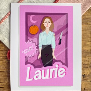 May include: An illustrated card in pink and purple featuring a cartoon character of a woman with brown hair, wearing a white shirt and black trousers. The character is holding a knife and a pumpkin. The card has the text "Laurie" and "brother sold separately" on it.