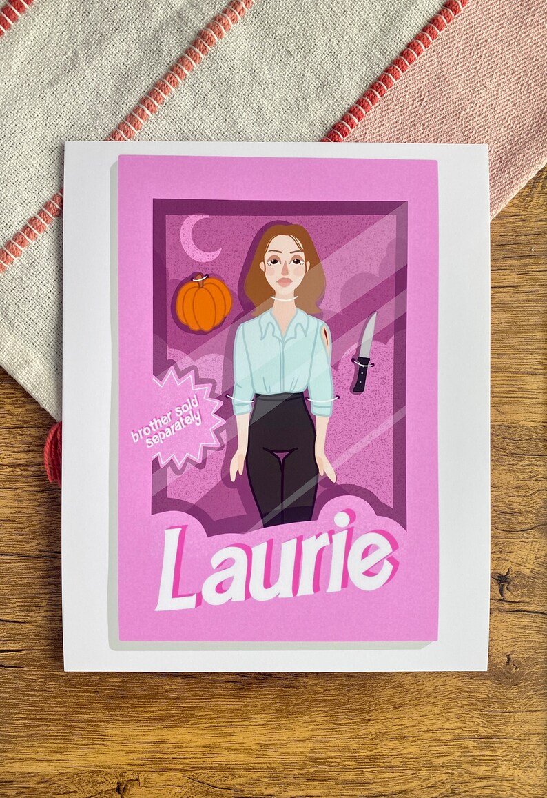 May include: An illustrated card in pink and purple featuring a cartoon character of a woman with brown hair, wearing a white shirt and black trousers. The character is holding a knife and a pumpkin. The card has the text "Laurie" and "brother sold separately" on it.