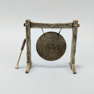 May include: A wooden gong stand with a round gong hanging from it. The gong is made of dark brown metal and has a textured surface. A wooden mallet is leaning against the stand.