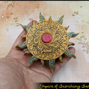 May include: A gold sun-shaped pendant with a turquoise border and a red jewel in the center. The pendant is on a hand, and the background is a light brown grid.