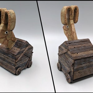 May include: A wooden box with wheels, painted to look like weathered wood, with a stitched, tan, rabbit head sticking out of the top. The rabbit has large ears and a pink nose.