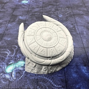 May include: White 3D printed tabletop game piece with a circular design featuring a central circle surrounded by concentric rings of symbols and numbers. The piece is shaped like a stylized wing or a crescent moon.