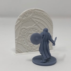 May include: A miniature figure of a person wearing a cloak and holding a shield and sword, standing in front of a white, 3D printed, circular, decorative wall panel.