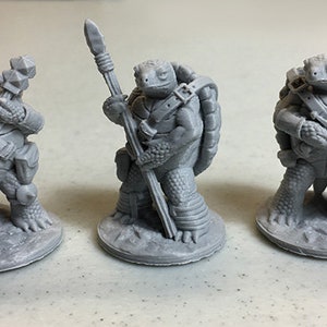 May include: Three grey 3D printed miniature turtle figurines. The turtle on the left is holding a hammer and shield. The turtle in the middle is holding a spear. The turtle on the right is holding a sword and shield.